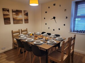 A dining room with a wooden table set for six, surrounded by chairs. A wall clock and three paintings are visible.