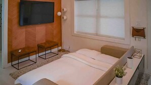 A modern bedroom with a large bed, a flat-screen TV mounted on the wall, a window with blinds, and a small bedside table with a plant.