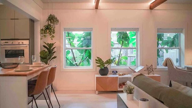 A modern kitchen with a dining area, a sofa, and large windows with greenery outside.