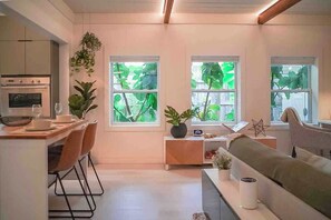 A modern kitchen with a dining area, a sofa, and large windows with greenery outside.