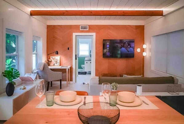 A modern living room with a dining area, a television mounted on the wall, and a door leading to another room.