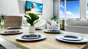 Un comedor con una mesa de madera dispuesta para cuatro personas, una planta en maceta y un cuadro con motivos playeros en la pared.