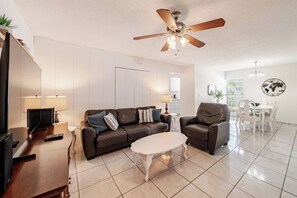 A living room with a brown leather sofa, a white coffee table, a ceiling fan, and a dining area with white chairs and a table.