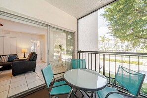 A balcony with a glass table and chairs, a sofa with cushions, and a view of a tree and buildings.