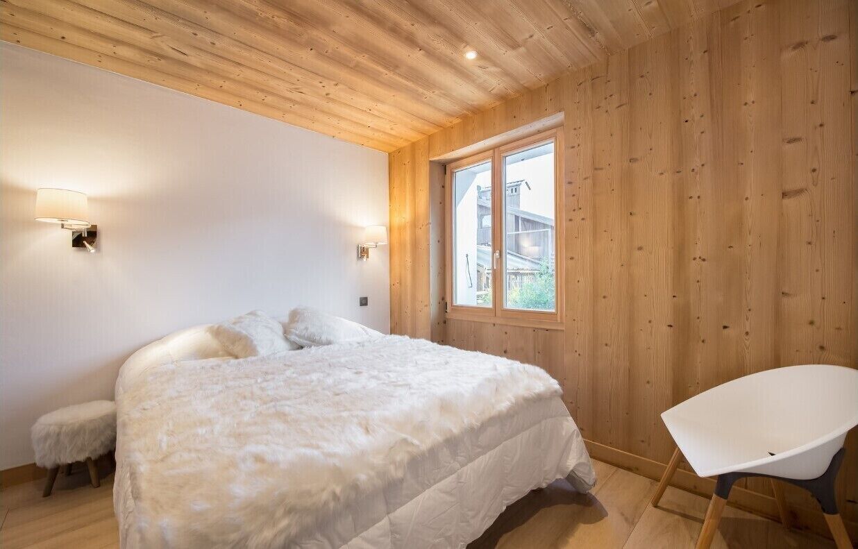 A bedroom with a large bed, a small stool, a white chair, wooden walls, and a window.
