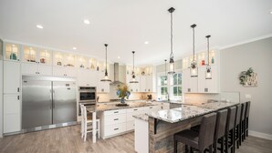 Een moderne keuken met witte kasten, een roestvrijstalen koelkast, een marmeren kookeiland met barkrukken, hanglampen en een groot raam.
