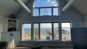 Een kamer met een groot erkerraam met uitzicht op zee, een plafond met zichtbare balken en een beeld van het strand en boten.