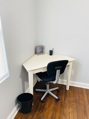 A corner office with a white desk, a black office chair, a small trash bin, and a motivational sign.