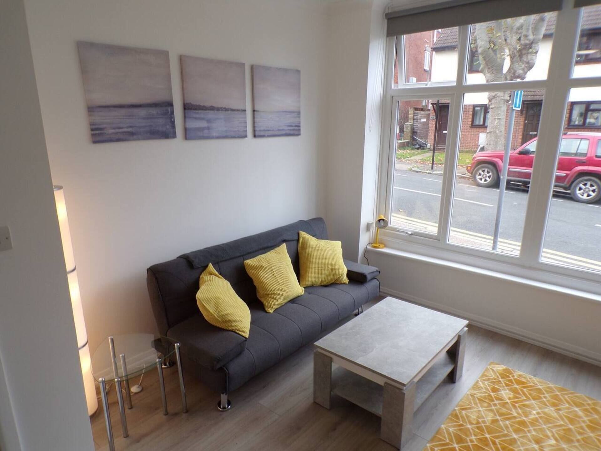 A modern living room with a grey sofa, two yellow cushions, a glass coffee table, and a view of a street with cars through a large window.
