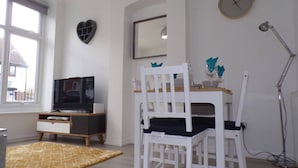 A modern living room with a television, a dining table, chairs, a heart-shaped wall decoration, a clock, and a lamp. - Inviting 3-bed Apartment in Southend-on-sea (Southend-on-Sea)