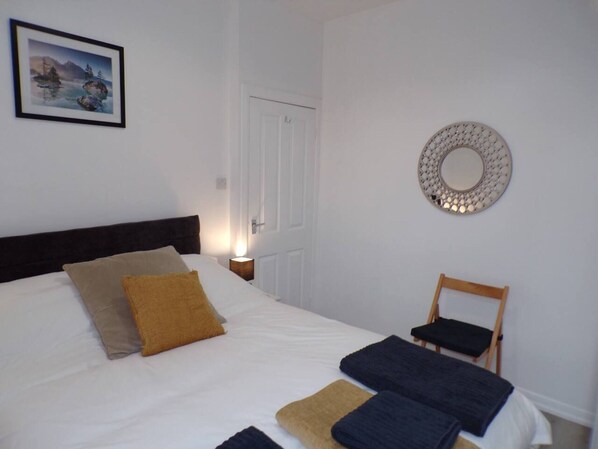 A neatly made bed with a headboard, a bedside table with a lamp, a chair, and a wall-mounted mirror. - Inviting 3-bed Apartment in Southend-on-sea (Southend-on-Sea)