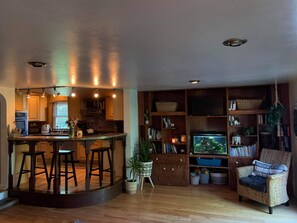 A kitchen with stools, a dining table, and a bar area. There is a bookshelf filled with books and decorative items.