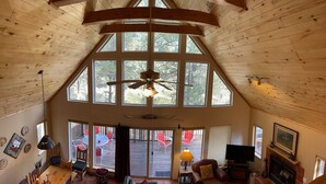 A spacious living area with a high ceiling, large windows, a ceiling fan, a fireplace, and a television. - Nestled in the pines on 2 acres, secluded, close-to-nature house. Pet-Friendly! (Angel Fire)