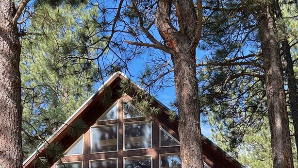 A cabin with a wooden exterior, large windows, and a balcony with red umbrellas. - Nestled in the pines on 2 acres, secluded, close-to-nature house. Pet-Friendly! (Angel Fire)