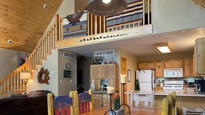 A spacious living area with a wooden dining table, colorful chairs, a staircase leading to a loft, and a kitchen with wooden cabinets and modern appliances. - Nestled in the pines on 2 acres, secluded, close-to-nature house. Pet-Friendly! (Angel Fire)
