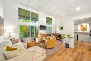 Een ruime woonkamer met een bank, rieten stoelen, een salontafel en een eettafel. Er is een keuken met witte kasten en een spoelbak. Grote ramen bieden uitzicht op groen buiten.