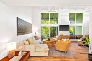 Un salon spacieux avec un grand canapé, des fauteuils en osier, un téléviseur à écran plat et une vue sur une végétation luxuriante grâce à de grandes fenêtres.