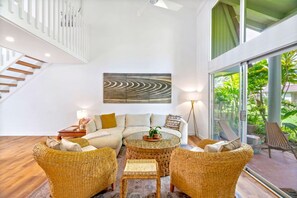 A spacious living room with a large sofa, two wicker chairs, a coffee table, and a floor lamp. A staircase is visible in the background, and a large window offers a view of greenery outside. - Puamana 8B · Puamana Repose (Princeville)