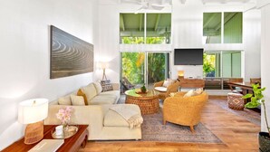 Ein geräumiges Wohnzimmer mit einem großen Sofa, Korbsesseln, einem Flachbildfernseher und Blick auf üppiges Grün durch große Fenster.