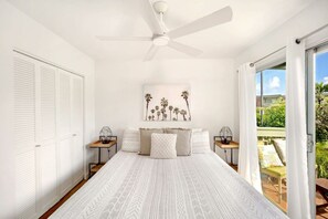 A bedroom with a large bed, a ceiling fan, a view of the outdoors, and a painting on the wall. - Puamana 8B · Puamana Repose (Princeville)