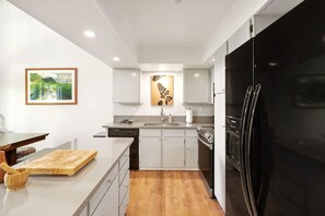 A modern kitchen with white cabinetry, a black refrigerator, and a wooden cutting board. - Puamana 8B · Puamana Repose (Princeville)