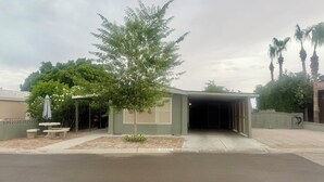 A single-story building with a covered garage entrance, a small patio with a table and chairs, and a tree in front. - Relaxing Foothills Home RV Hookup and Parking (Yuma)