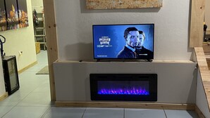 Ett modernt vardagsrum med en platt-TV, en öppen spis och en tavla som föreställer en stadsbild.