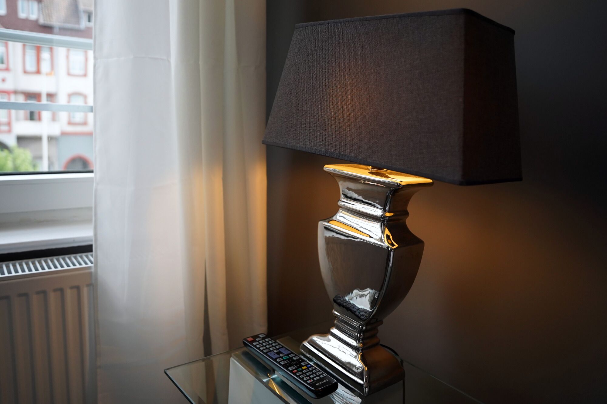 En modern lampa med en metallbas och en svart lampskärm, placerad på ett glasbord bredvid ett fönster med utsikt över byggnader.