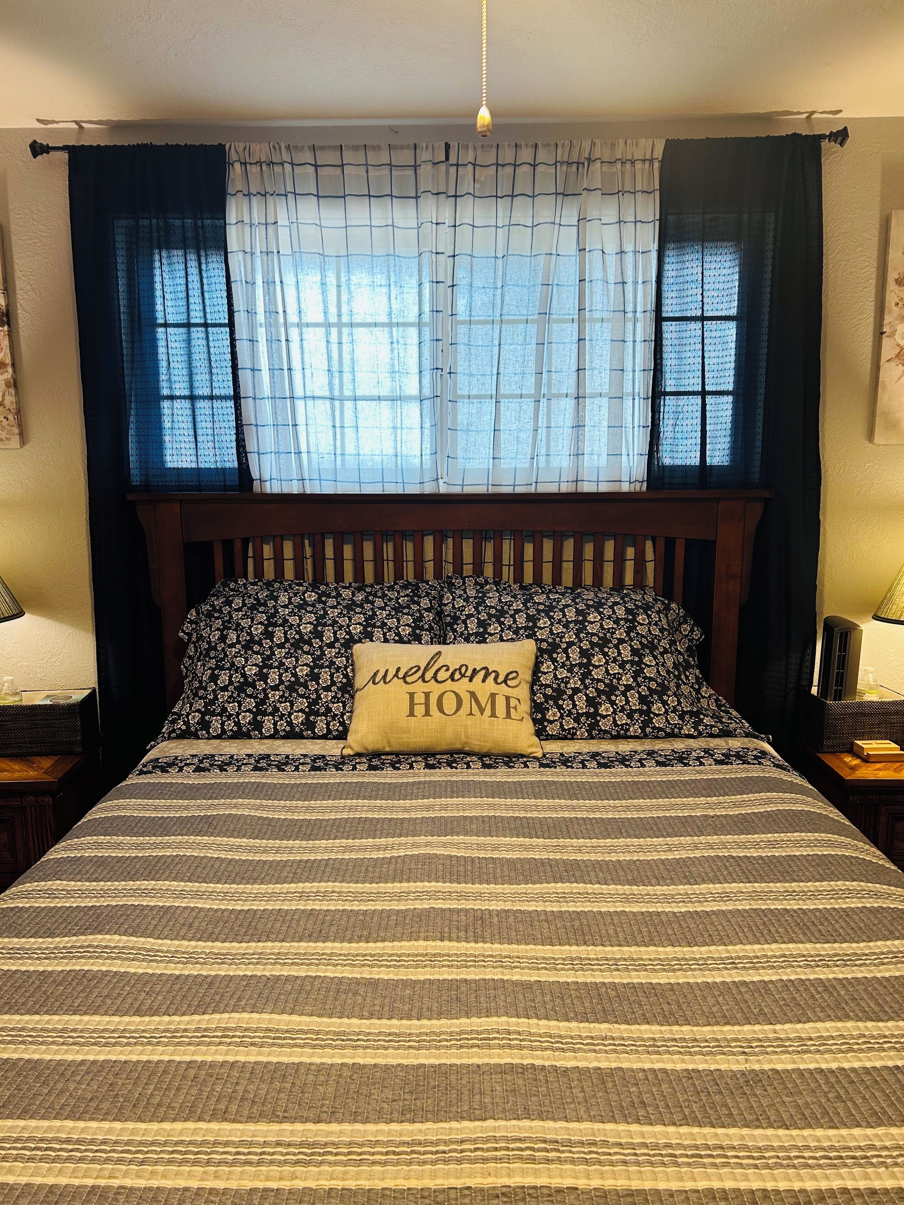 Uma cama com colcha listrada, um travesseiro com a inscrição 'welcome HOME' e uma janela com cortinas leves.
