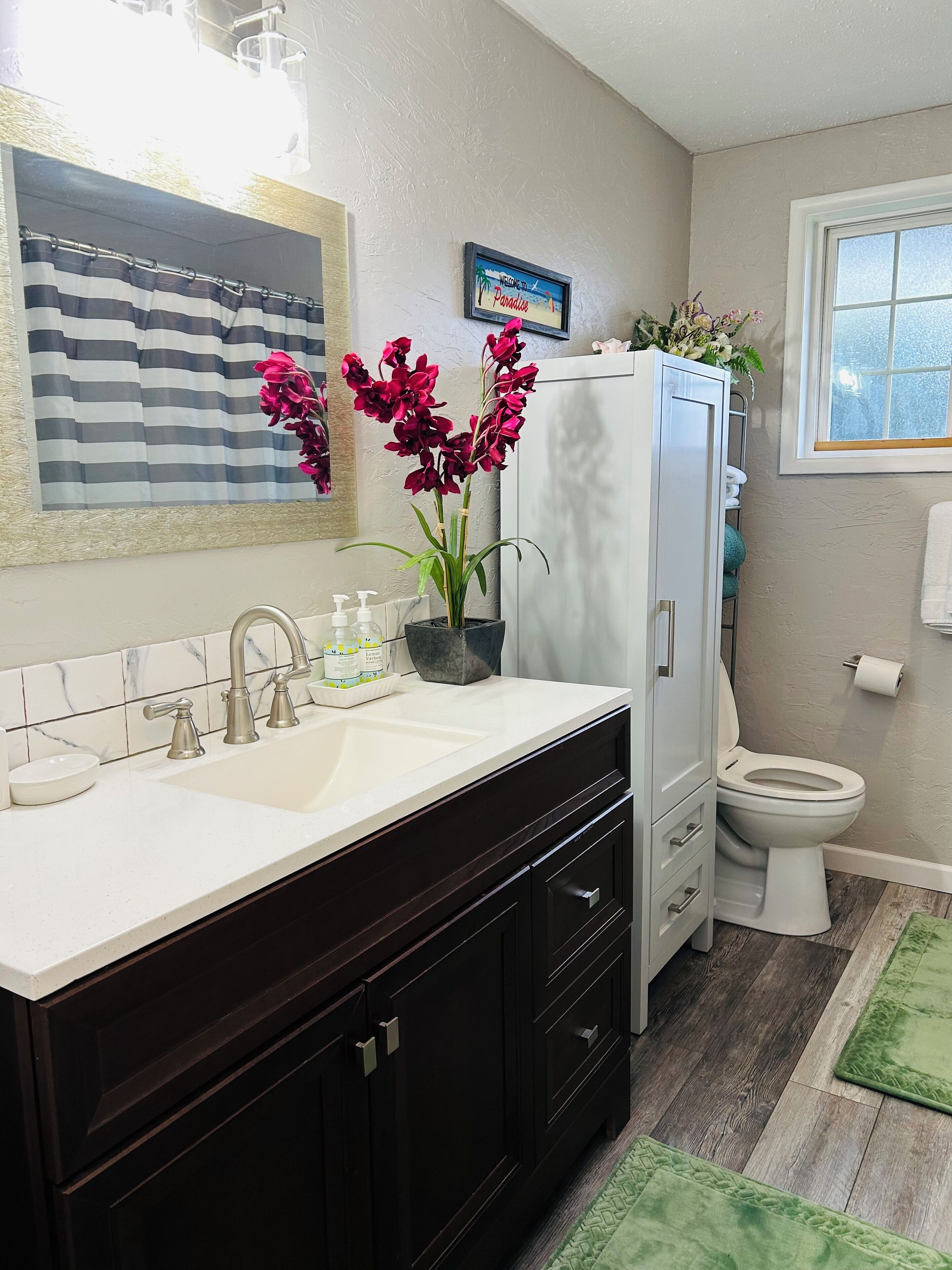 A bathroom with a white sink, dark cabinets, a toilet, and a green rug.
