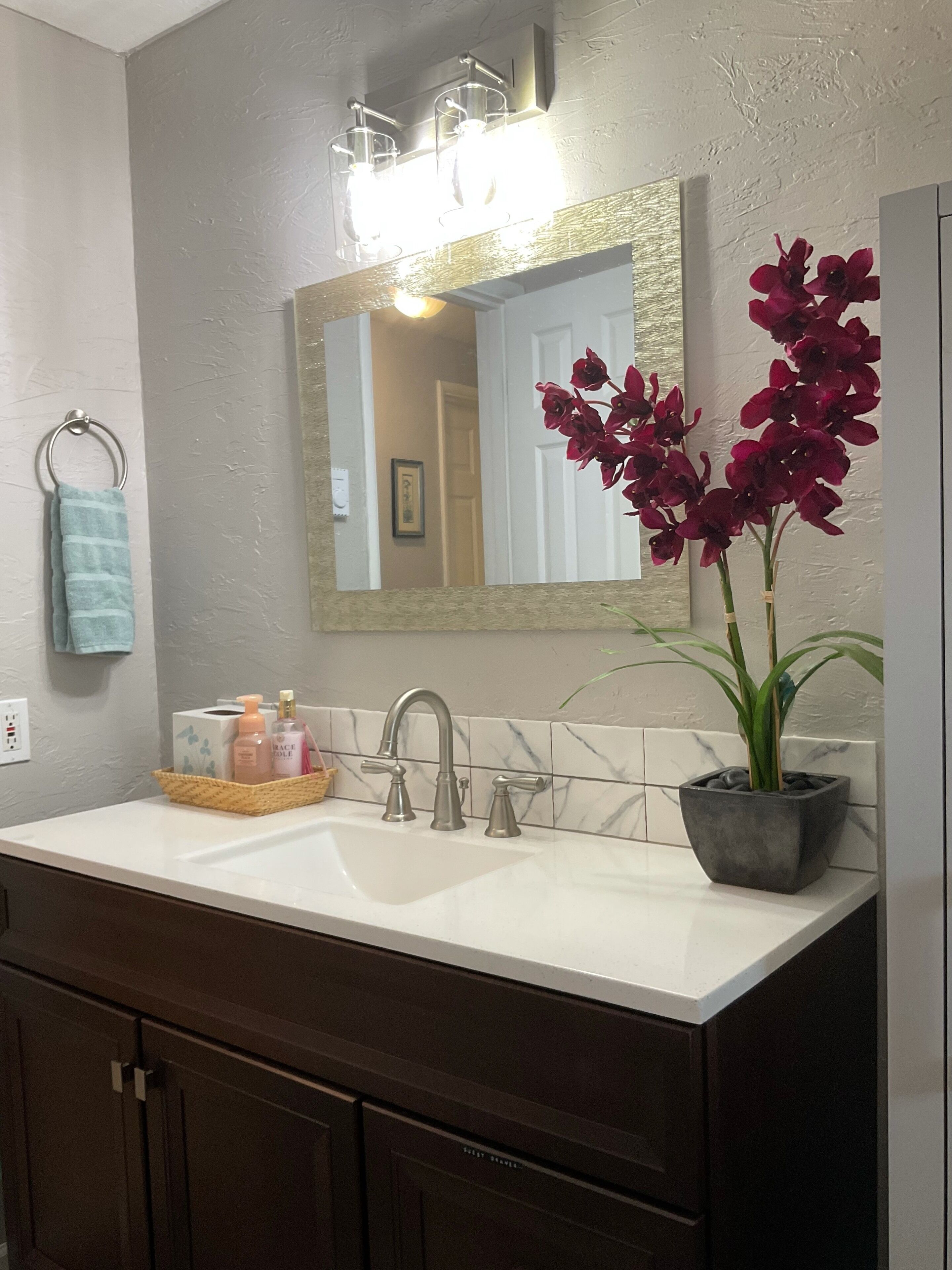 A bathroom with a white sink, a mirror, a towel rack, a potted plant, and toiletries on the counter.