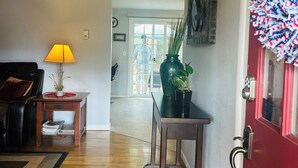 Ein Wohnzimmer mit Holzboden, einer schwarzen Ledersofa, einer Lampe, einem kleinen Tisch mit Pflanze und einer roten Tür mit Überwachungskamera.