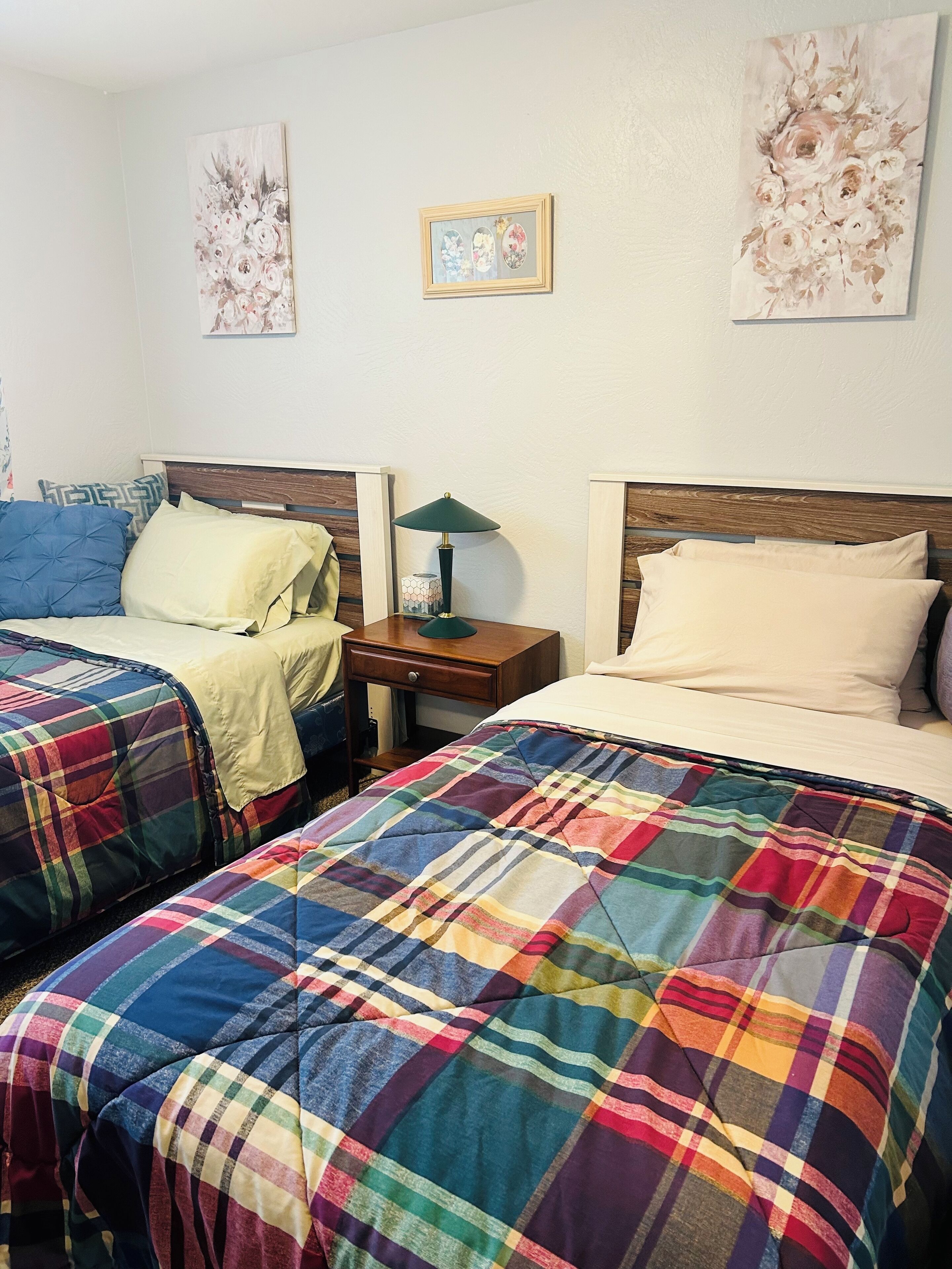 A bedroom with two beds, each with a colorful plaid blanket, a nightstand with a lamp, and floral artwork on the walls.
