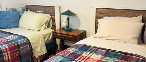 A bedroom with two beds, each with a colorful plaid blanket, a nightstand with a lamp, and floral artwork on the walls.