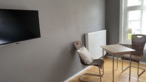 Un salon moderne doté d’un téléviseur à écran plat, d’une table à manger, de deux fauteuils en osier, d’un radiateur et d’une fenêtre laissant entrer la lumière naturelle.