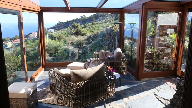 A sunlit room with a view of a hillside, wicker furniture, and a stone floor.