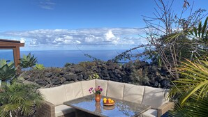 A patio with wicker furniture and a glass-top table, overlooking a rocky coastline and the sea.