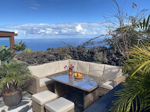 Um pátio com mobiliário de vime e uma mesa com tampo de vidro, com vista para um litoral rochoso e o mar.