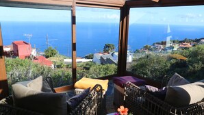 A balcony with wicker furniture, a small table with a vase of flowers, and a view of the sea and houses.