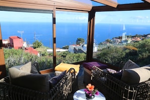 Varanda com móveis de vime, uma pequena mesa com um vaso de flores e vista para o mar e casas.
