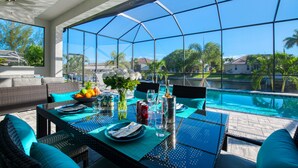 Un espace repas délimité par une paroi de verre, doté de mobilier d’extérieur et offrant une vue sur une piscine et des palmiers.
