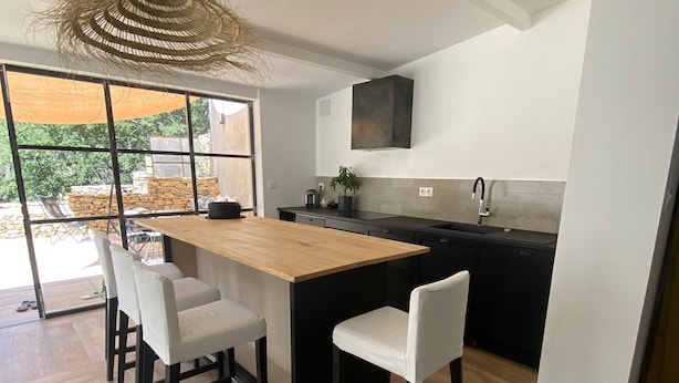 Une cuisine moderne dotée d’un îlot central, de meubles de rangement noirs et d’une grande fenêtre donnant sur un jardin.