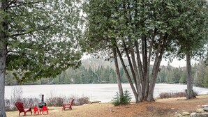 A lakeside setting with red Adirondack chairs, a stone fire pit, and a view of a calm lake surrounded by trees. - Magical Moments Hot tub & Swimmable Lake (Amherst)