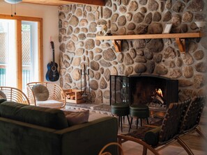 A cozy living room with a stone wall, a fireplace, wicker furniture, and a guitar. - Magical Moments Hot tub & Swimmable Lake (Amherst)