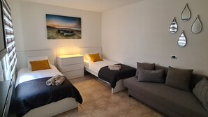 A bedroom with two beds, a sofa, a nightstand with a lamp, a painting, and wall mirrors. - Modern Studio in Rural Tinajo with Patio, BBQ & Coastal Views – Perfect Getaway! (La Costa)