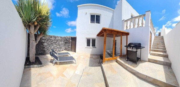 A modern villa with a stone wall, a wooden pergola, a barbecue area, and a staircase leading to the upper floor. - Modern Studio in Rural Tinajo with Patio, BBQ & Coastal Views – Perfect Getaway! (La Costa)