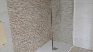 A modern bathroom with a glass shower enclosure, a textured wall, and a small rug on the floor. - Modern Studio in Rural Tinajo with Patio, BBQ & Coastal Views – Perfect Getaway! (La Costa)