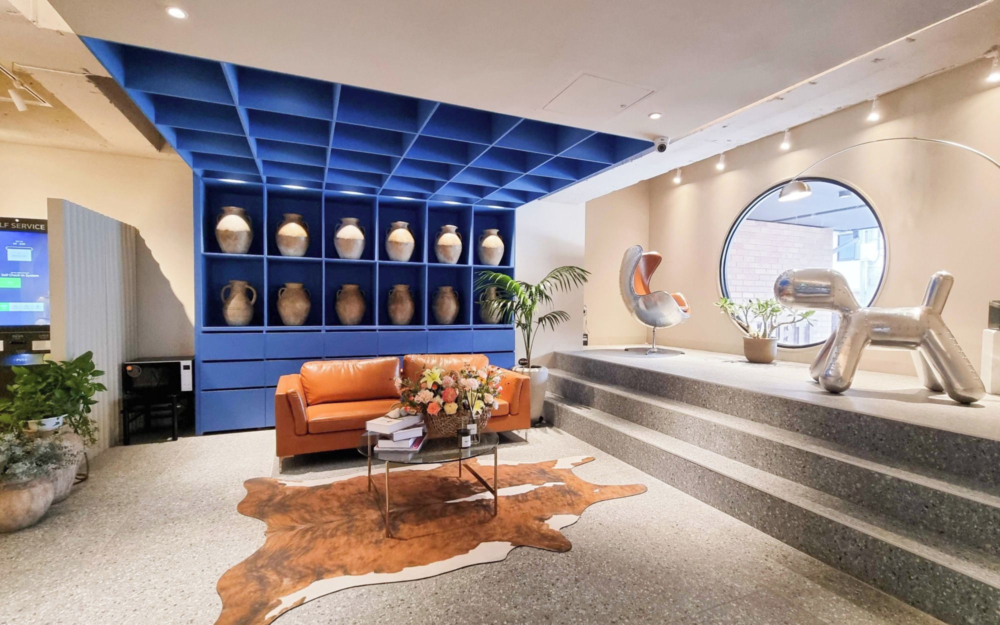 A modern lobby with a blue shelving unit displaying vases, a leather sofa, a cowhide rug, and a staircase with a round mirror.