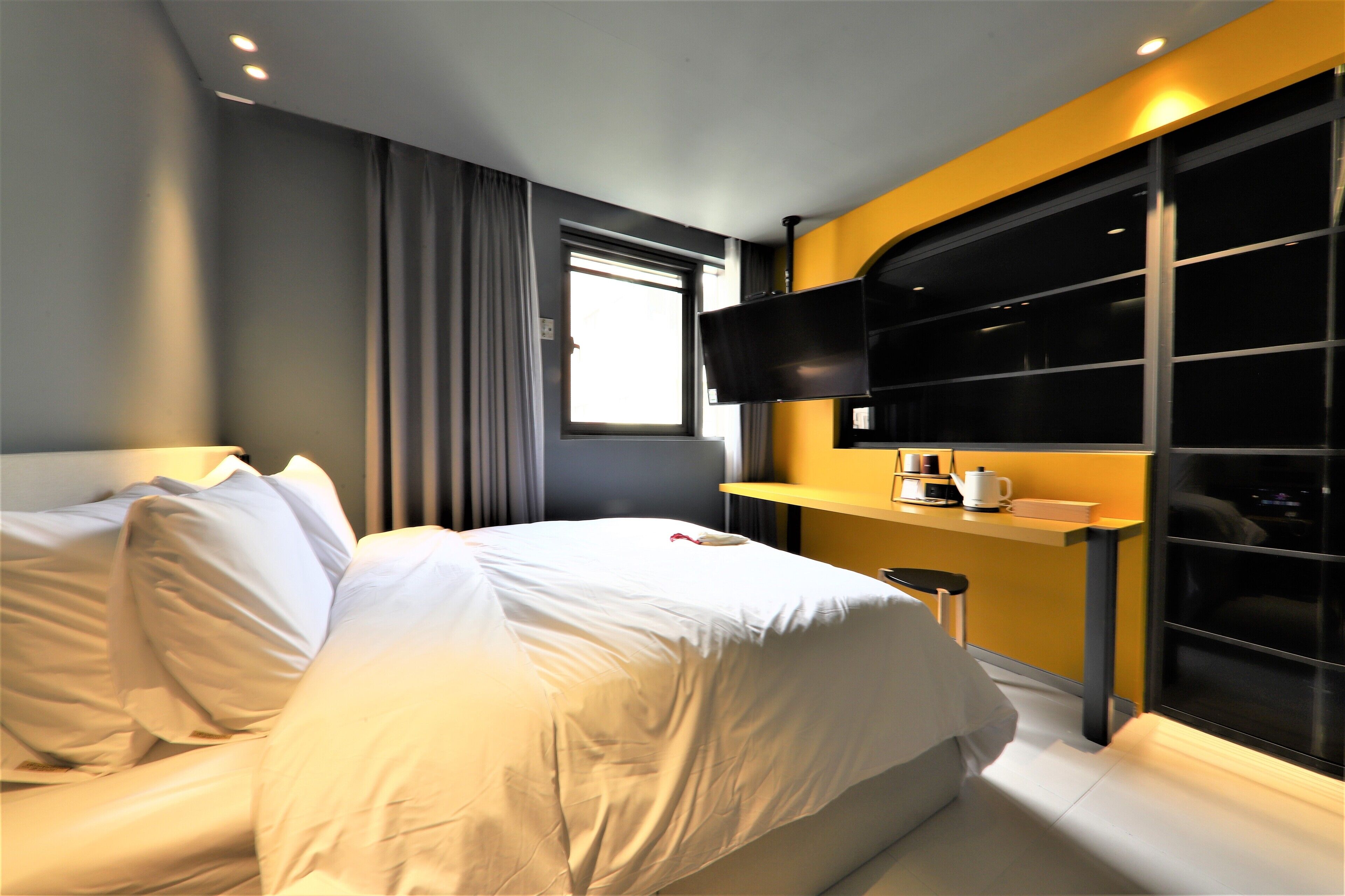 Una habitación de hotel moderna con una cama bien tendida, un televisor de pantalla plana montado en la pared, una mesita con cafetera y un ventanal con cortinas.