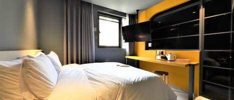 Una habitación de hotel moderna con una cama bien tendida, un televisor de pantalla plana montado en la pared, una mesita con cafetera y un ventanal con cortinas.